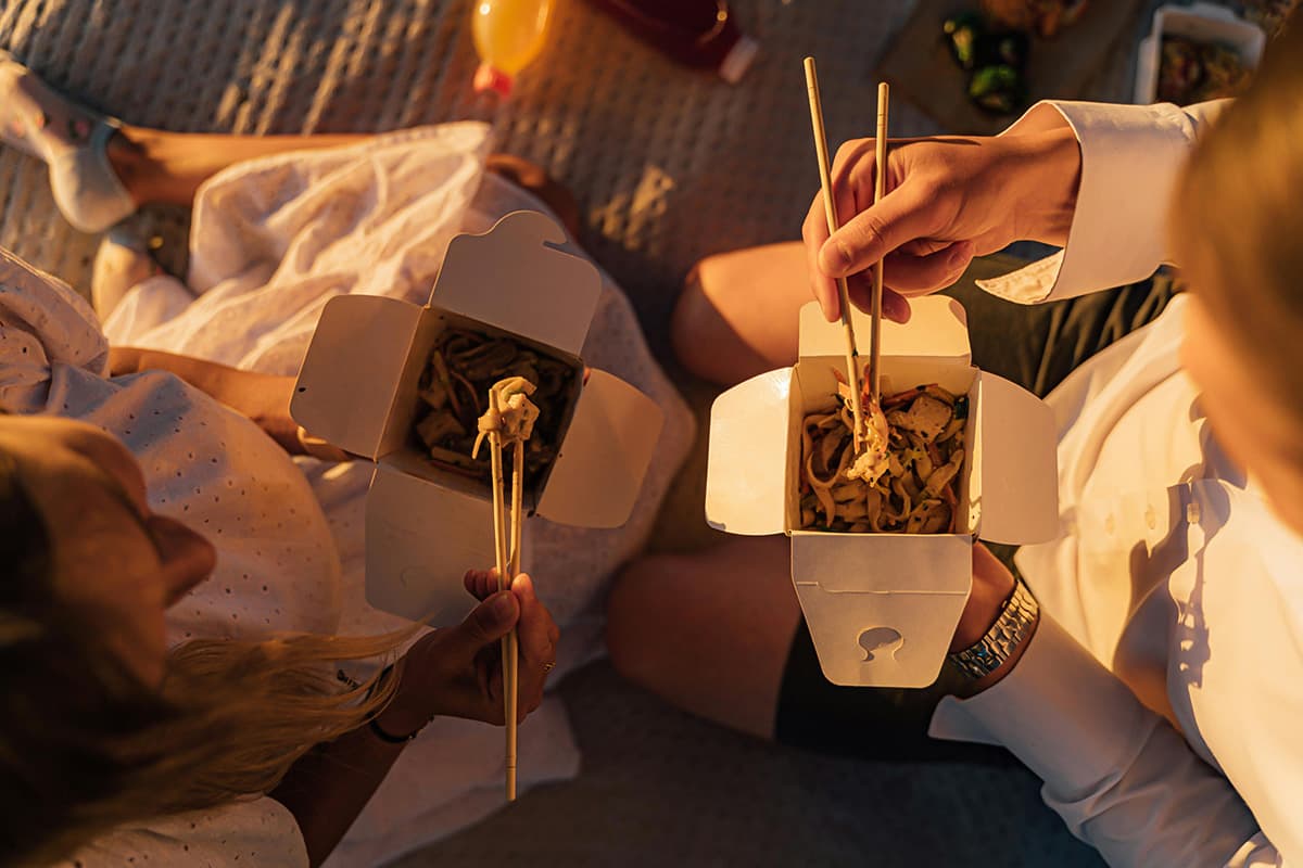 Two people enjoying a takeaway meal outdoors with white cardboard containers, eating noodles with chopsticks in warm golden light